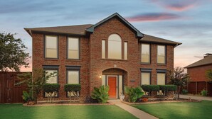 A two-story brick house with a front entrance, a well-maintained lawn, and a wooden fence. - Vista Valley Vacay, 5-bd w Pool & Cinema (Lewisville)