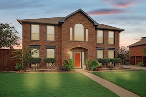 A two-story brick house with a front entrance, a well-maintained lawn, and a wooden fence. - Vista Valley Vacay, 5-bd w Pool & Cinema (Lewisville)