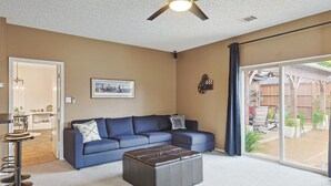 A living room with a blue sofa, a dark ottoman, a ceiling fan, and a large sliding glass door leading to an outdoor area. - Vista Valley Vacay, 5-bd w Pool & Cinema (Lewisville)
