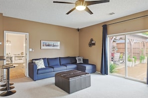 A living room with a blue sofa, a dark ottoman, a ceiling fan, and a large sliding glass door leading to an outdoor area. - Vista Valley Vacay, 5-bd w Pool & Cinema (Lewisville)
