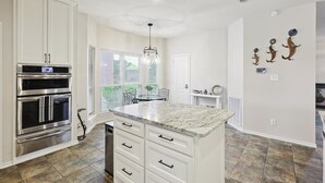 A kitchen with white cabinets, a double oven, a marble island, and a dining area with a table and chairs. - Vista Valley Vacay, 5-bd w Pool & Cinema (Lewisville)
