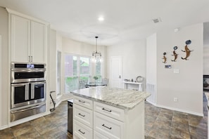 A kitchen with white cabinets, a double oven, a marble island, and a dining area with a table and chairs. - Vista Valley Vacay, 5-bd w Pool & Cinema (Lewisville)