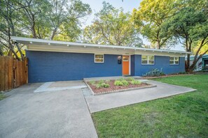 Una casa de un piso con fachada azul, puerta principal y pequeño jardín delantero con sendero de concreto y macizos de flores.