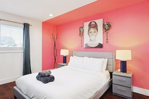 Ein Hotelzimmer mit einem Bett, zwei Nachttischen mit Lampen, einem Fenster mit Vorhängen und einer rosa Wand mit einem Gemälde.