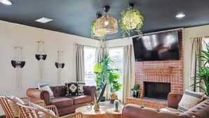 A living room with a brick fireplace, a large flat-screen TV, wicker furniture, and hanging plants. - Luxury 5BR Getaway w/ Pool, Hot Tub & cinema (DeSoto)