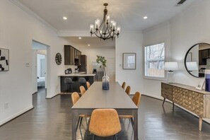 Un espace repas avec une longue table, des chaises, un lustre, une cuisine en arrière-plan et un buffet agrémenté d’un miroir.