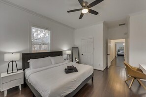 Une chambre à coucher comprenant un lit, des tables de chevet, un ventilateur de plafond, une fenêtre avec des stores, une porte et un couloir menant à une autre pièce.