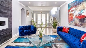 A modern living room with a glass coffee table, two blue sofas, a large abstract painting, and a staircase leading to another floor. - Luxury Tropical Living - 4BR w Backyard Pool Oasis (Miami Lakes)