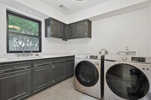A modern laundry room with built-in washers and dryers, a large window, and dark cabinetry. - Best of Carrollton - Luxury, Pool, Hot Tub, Games (Carrollton)