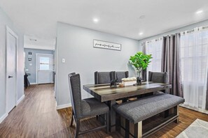 A dining area with a wooden table, grey upholstered chairs, and a bench. There is a plant on the table and a wall with a framed quote. - 4 Bedrooms, Brick Retreat With Gameroom & Backyard (Grand Prairie)