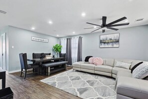 A modern living room with a dining area, a ceiling fan, and a large window with curtains. - 4 Bedrooms, Brick Retreat With Gameroom & Backyard (Grand Prairie)