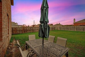 A patio with a table and chairs, a green umbrella, and a wooden fence in the background. - 4 Bedrooms, Brick Retreat With Gameroom & Backyard (Grand Prairie)