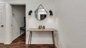 A hallway with a white door, a wooden floor, a small table with plants, and a round mirror with two wall-mounted lights. - Modern and Luxury DFW Mansion With Backyard Oasis (Fort Worth)