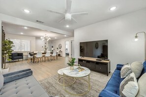 A modern living room with a sofa, a coffee table, a TV, and a dining area with chairs and a table. - Luxury 4bed/2bath Hidden Gem w/ Backyard Oasis (North Richland Hills)