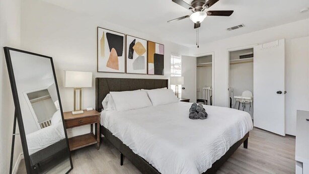 A modern bedroom with a bed, a ceiling fan, a bedside table with a lamp, a mirror, and abstract wall art. - Luxury 4bed/2bath Hidden Gem w/ Backyard Oasis (North Richland Hills)