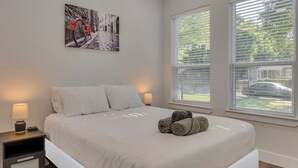 Een slaapkamer met een bed, twee nachtkastjes, een lamp, een schilderij en twee ramen met zonwering.