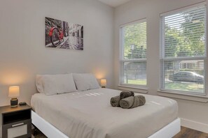 Ein Schlafzimmer mit einem Bett, zwei Nachttischen mit Lampen, einem Fenster mit Jalousien und einem Wandbild.