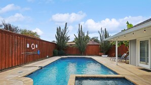 A rectangular swimming pool with a wooden deck surrounding it, a covered patio area with chairs, and a fenced backyard. - Best of Allen at Fountain Park 5-bd/2-ba w/ Pool (Allen)