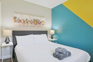 A hotel room with a bed, two bedside tables with lamps, a wall painting, and a blue and yellow accent wall. - Best of Allen at Fountain Park 5-bd/2-ba w/ Pool (Allen)