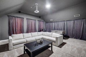 A living room with a sectional sofa, a coffee table, and a ceiling-mounted projector. - Stylish, Spacious, Convenient - 6-bd/2.5-ba + Pool (Fort Worth)