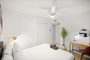 Ein Schlafzimmer mit Bett, Schreibtisch, Stuhl, Deckenventilator, einer Zimmerpflanze und einem Spiegel.