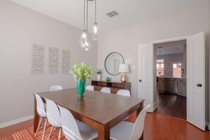 Een eetruimte met een houten tafel, witte stoelen, een groene vaas met bloemen, hanglampen, en een keuken zichtbaar door een open deur.