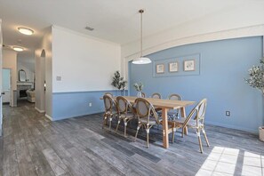 A dining area with a wooden table and chairs, a hanging light fixture, and framed artwork on the wall. - The Blue Holly - Bright & Spacious With a Pool (Denton)