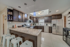 A modern kitchen with dark cabinets, a granite island, white bar stools, and pendant lighting. - Central Plano 4-bd/2-ba Pool Retreat, Sleeps 10 (Plano)