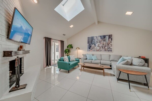A spacious living room with a fireplace, a large flat-screen TV, a sofa set, and a cityscape painting. - Central Plano 4-bd/2-ba Pool Retreat, Sleeps 10 (Plano)