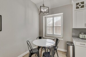 Uma área de refeições com uma mesa redonda, cadeiras pretas, um lustre pendente, uma janela com persianas e uma cozinha com uma torradeira.