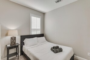 A bedroom with a bed, a nightstand, a lamp, and a window with blinds. - Trendy and Modern Home w/ a Private Pool (Rowlett)