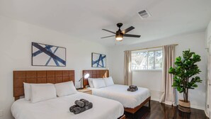 Ett hotellrum med två sängar, en takfläkt, konstverk på väggarna, ett stort fönster med gardiner och en krukväxt.