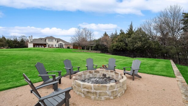 A stone fire pit with four Adirondack chairs, a well-manicured lawn, and a large house in the background. - Lakefront Mansion With Pool and Cinema (Wylie)