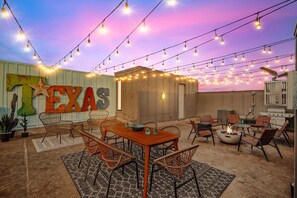 Eine gemütliche Terrasse im Freien mit Lichterketten, einem Holztisch, Korbstühlen, einer Feuerstelle und einer Wand im Texas-Stil.