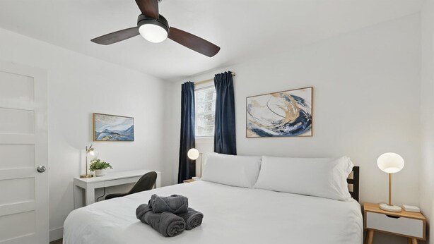 Habitación de hotel con cama, escritorio, silla, ventilador de techo, obras de arte y mesita de noche con lámpara.