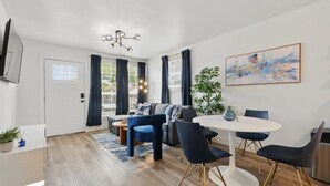 Una sala de estar moderna con un sofá, una mesa de comedor y sillas, un televisor de pantalla plana montado en la pared y un cuadro abstracto grande.