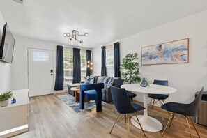 A modern living room with a sofa, dining table, and chairs, a flat-screen TV mounted on the wall, and a large abstract painting. - Charming Historic 3B Heart of Dallas - Sleeps 12 (Dallas)
