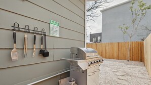 Una parrilla para barbacoa con un tanque de gas, un juego de utensilios colgados en una pared y un cartel con normas para la barbacoa.