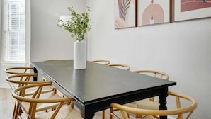 Een eetgedeelte met een zwarte tafel, rieten stoelen, een witte vaas met een plant en abstracte wanddecoratie.