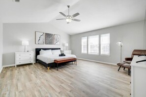 Ein Schlafzimmer mit einem Bett, Nachttischen, einem Schreibtisch, einem Sessel und großen Fenstern, sowie einem Deckenventilator.