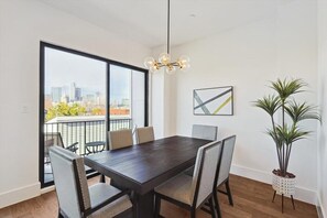 A dining area with a dark wooden table, six chairs, a hanging light fixture, a potted plant, and a city view through large windows. - 3-bd Luxury Living w Rooftop Garden Views (Dallas)