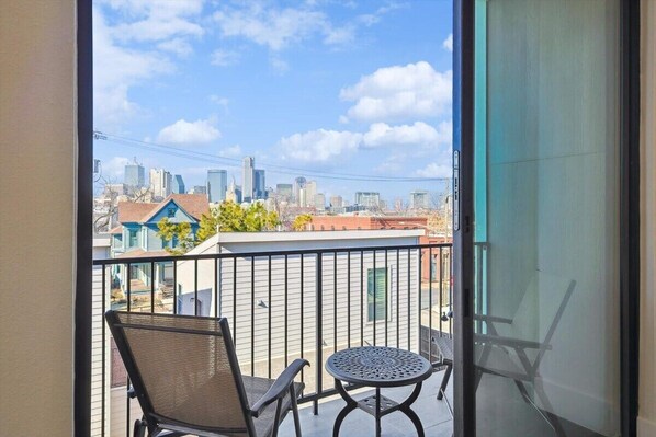 Ein Balkon mit Stuhl und Tisch, mit Blick auf eine Stadtlandschaft aus Gebäuden und einem klaren Himmel.