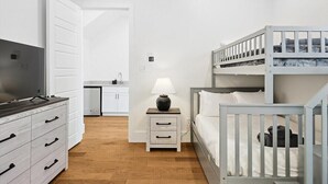 Et soveværelse med køjeseng, et natbord med lampe, et fjernsyn på en kommode og et tekøkken i baggrunden.