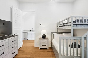 Ein Schlafzimmer mit Etagenbett, Nachttisch mit Lampe, Fernseher auf einer Kommode und einer Küchenzeile im Hintergrund.