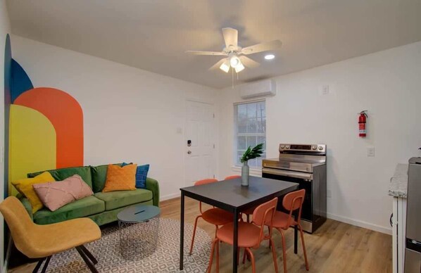 A compact living space with a green sofa, orange chairs, a small dining table, a ceiling fan, and a kitchen area with an oven. - Heart of Houston - 2-bd/1-ba With Grill (Houston)