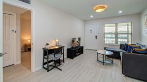 A modern living room with a sofa, a TV on a stand, a small table, and a window with blinds. - Greater Uptown 1bd, 1BA Apt, Mins to Attractions (Houston)
