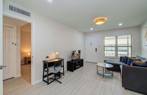 A modern living room with a sofa, a TV on a stand, a small table, and a window with blinds. - Greater Uptown 1bd, 1BA Apt, Mins to Attractions (Houston)