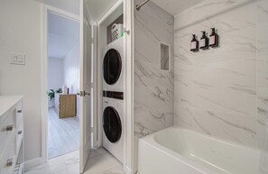 Et moderne badeværelse med væg i hvid marmor, indbygget vaskemaskine og tørretumbler, et badekar og tre toiletartikler monteret på væggen.