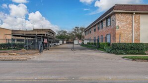 A gated parking area with a brick building and a covered structure in the background. - Greater Uptown 1bd, 1BA Apt, Mins to Attractions (Houston)