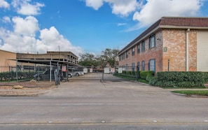 A gated parking area with a brick building and a covered structure in the background. - Greater Uptown 1bd, 1BA Apt, Mins to Attractions (Houston)